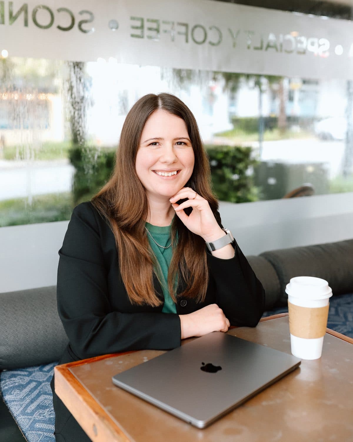 Jen Ross, founder of Mercury Jane, smiling with her chin on her hand at a coffee shop table, laptop and coffee beside her